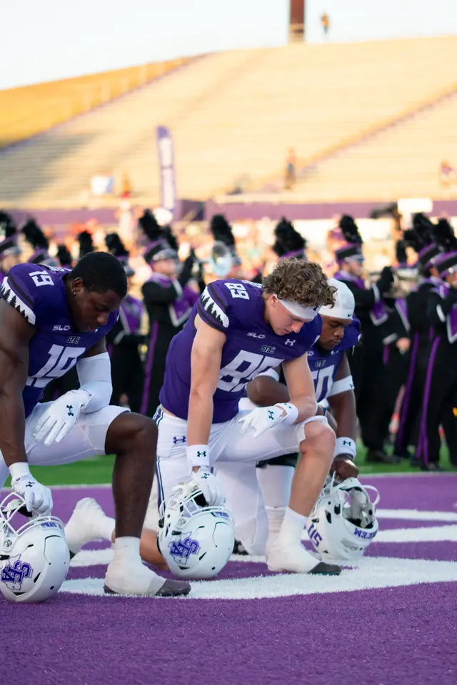 Three college football players in purple jerseys kneeling in the end zone before kickoff, marching band behind them